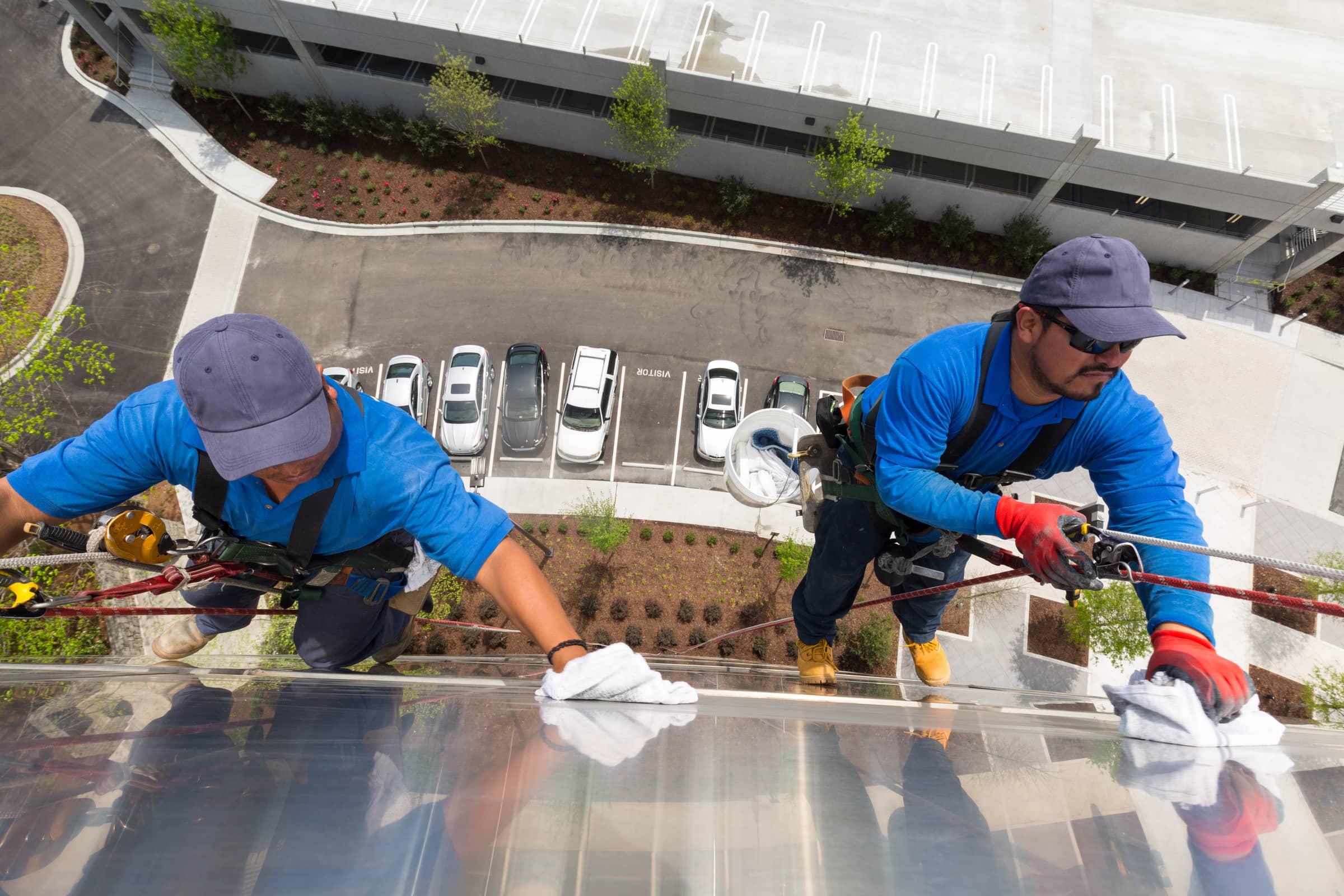 Two men cleaning windows on office building attached by ropes. Picture is taken looking downwards towards the car park below